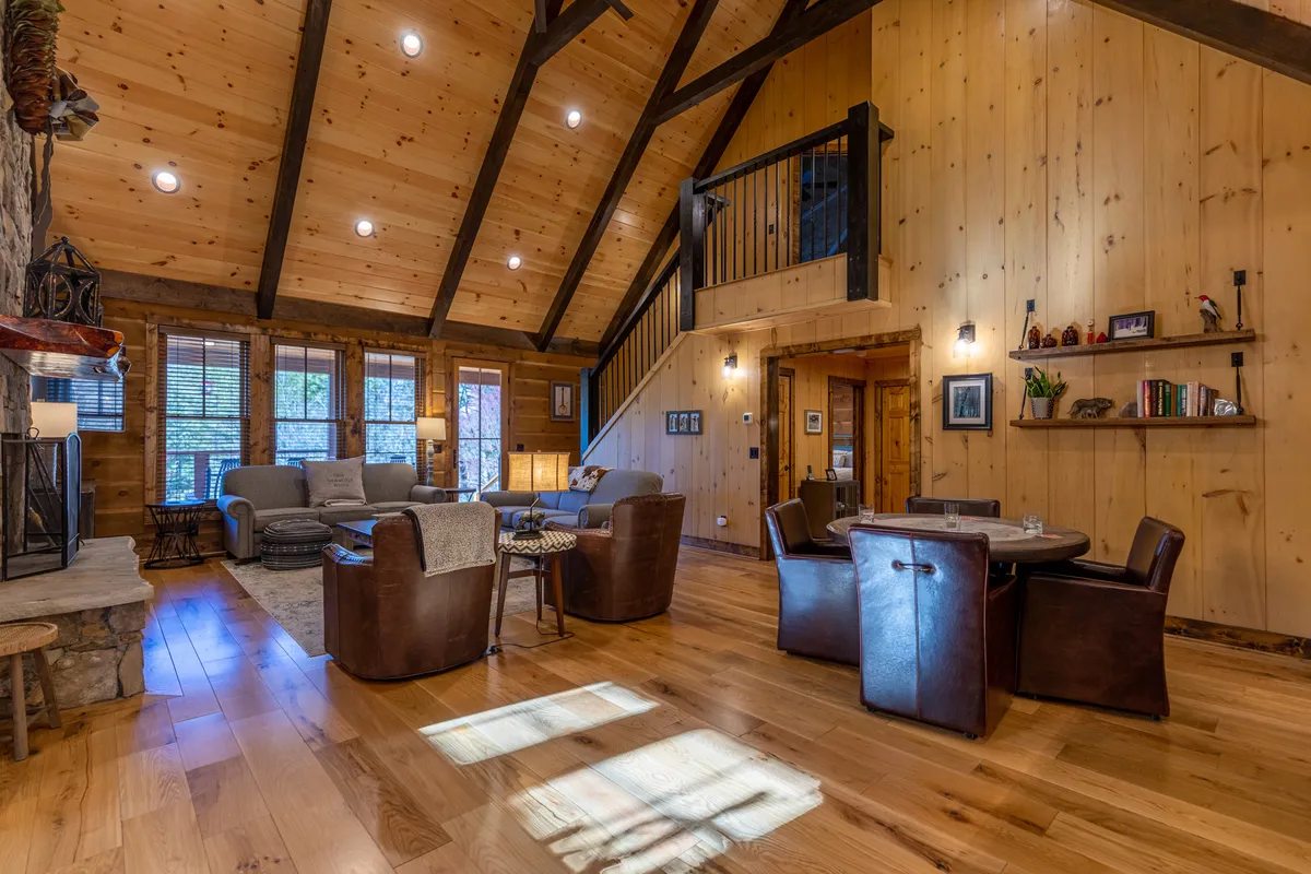 This image captures an open-concept living area with a stone fireplace, exposed wooden beams, large windows streaming in sunlight, a comfortable seating area, and a dining table in the background. The warm, welcoming atmosphere highlights spaciousness and natural light.