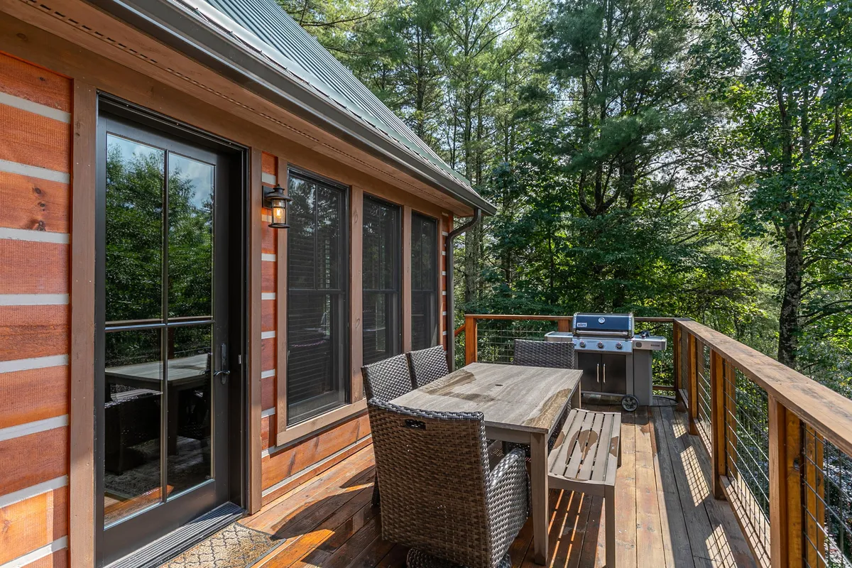 A wooden deck surrounded by trees, featuring a large outdoor dining table and a gas grill. The area is set for socializing, outdoor meals, and grilling, with ample space for gatherings against a backdrop of natural greenery.