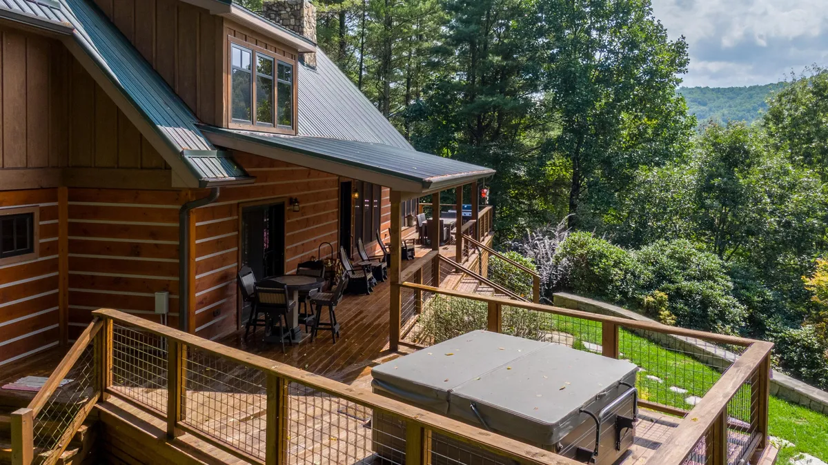 A spacious wooden deck is featured, with a bubbling hot tub prominently placed for soaking, surrounded by mature trees and adjacent to the house’s exterior wall. The scene exudes relaxation, with seating options available and a sense of privacy provided by the lush greenery.