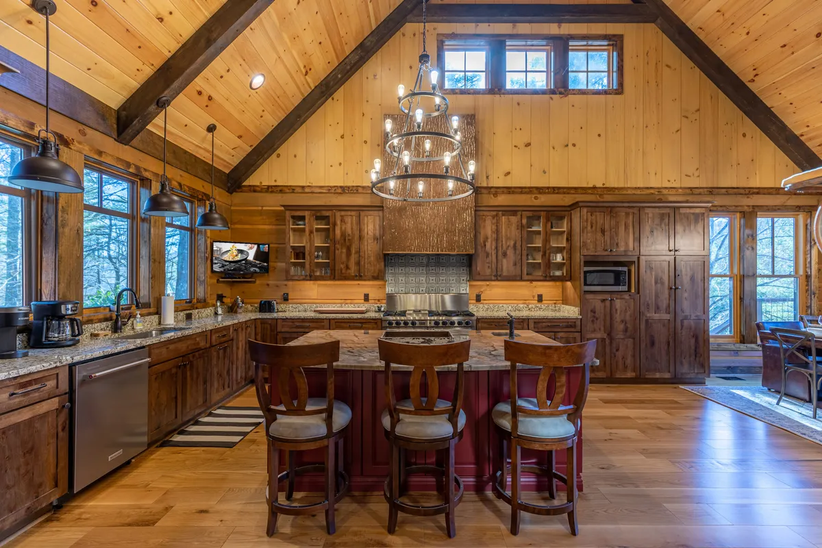 A bright kitchen scene with a large island and seating, elegant chandelier, granite countertops, wooden cabinetry, and wide windows letting in sunshine. The layout encourages both cooking and casual socializing.