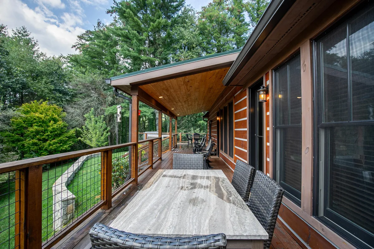 This image shows a wooden outdoor dining table surrounded by woven chairs, with lush green foliage and a sturdy wooden railing in the background. The table is set in an inviting, tranquil spot that is clearly meant for shared meals, sunset dinners, or socializing outdoors.