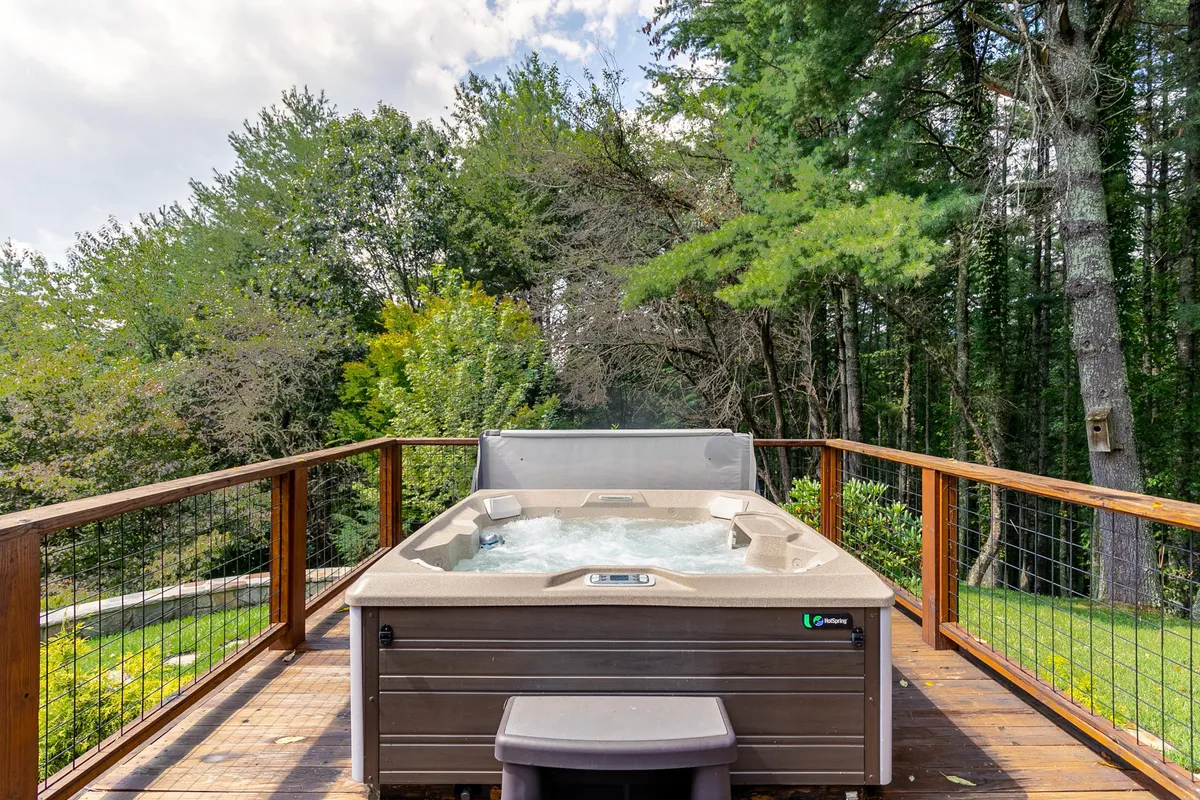 A spacious wooden deck featuring a hot tub, set against a backdrop of tall trees and greenery. The hot tub area is positioned for privacy and relaxation, making it ideal for unwinding under the stars after a day of adventure.