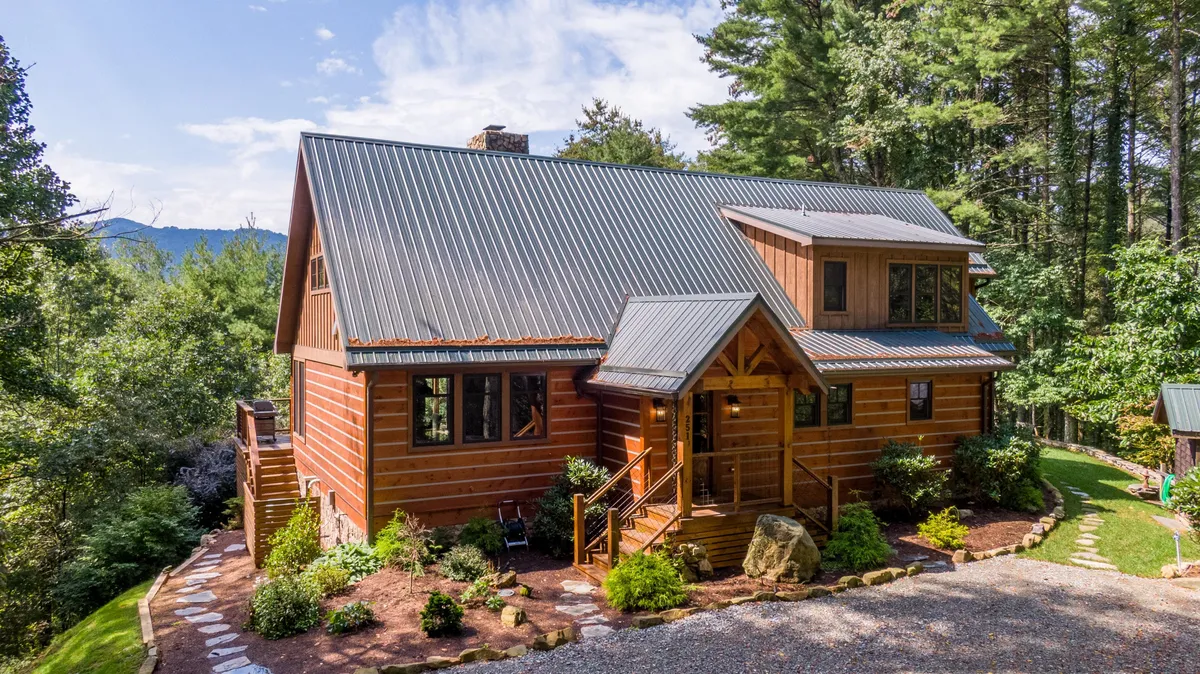 The image showcases a charming luxury log cabin nestled among lush greenery in Banner Elk, North Carolina. The spacious front porch invites relaxation, with sweeping mountain views perfect for sunset watching. The scene exudes rustic tranquility, with hints of a cozy atmosphere ideal for unwinding, sipping morning coffee, or gathering for campfire evenings.