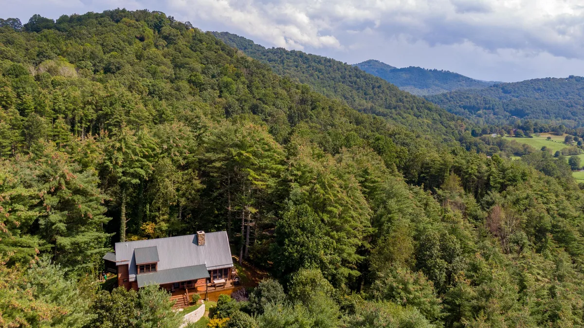 This image shows the house surrounded by tall trees and a scenic mountain backdrop. The porch overlooks a peaceful landscape that changes with the seasons, offering opportunities for sunrise views and stargazing at night. The lush greenery suggests proximity to hiking trails and natural exploration.