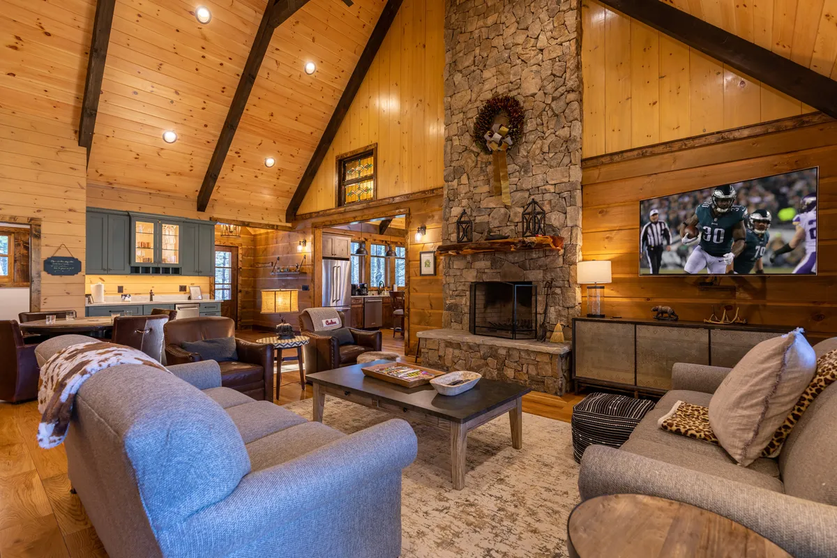 A living room scene centered around a striking stone fireplace, wood beams, comfortable seating, and a large TV. The space is designed for relaxing, movie nights, or socializing with family and friends in a warm, welcoming setting.