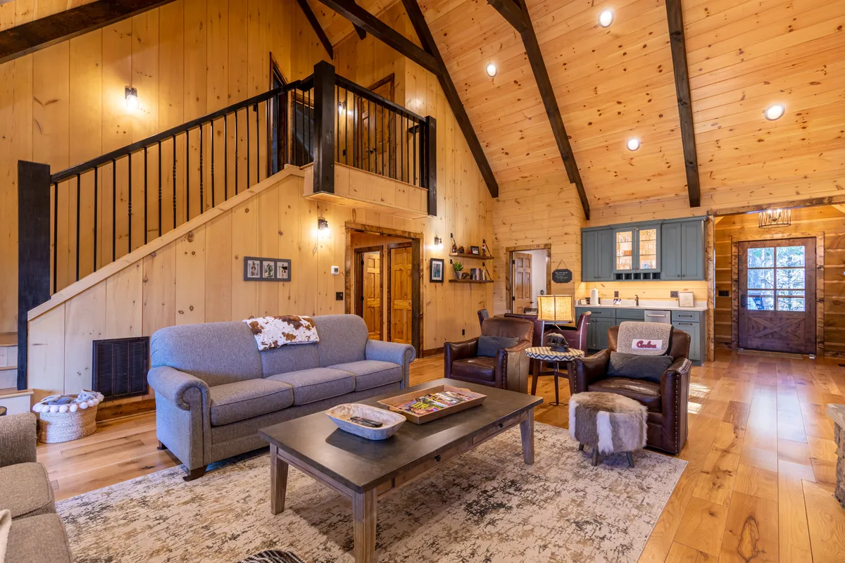 A wide-angle view of a spacious, airy living area featuring large wooden beams, a plush sofa, a visible wooden staircase, blue kitchen cabinets, and tall windows that invite abundant natural light. The layout visually links the living room, kitchen, and dining area.
