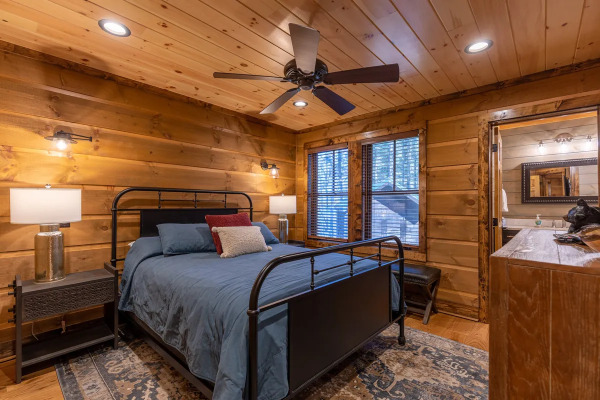 A welcoming queen bedroom with blue bedding, wooden walls, nightstands with lamps, and large windows with blinds. The room feels both cozy and spacious, perfect for unwinding after a day in the mountains.