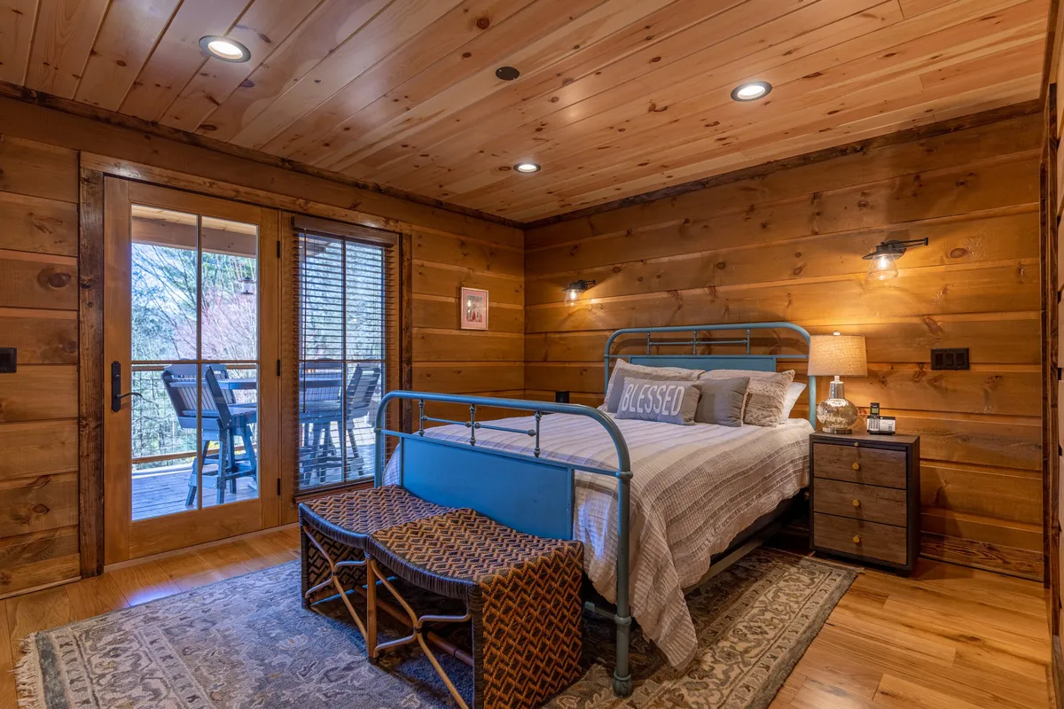 A tranquil queen bedroom bathed in natural light, featuring a cozy bed with plush linens, a woven bench at the foot, large windows with mountain views, a nightstand, and wall art that adds a touch of warmth.