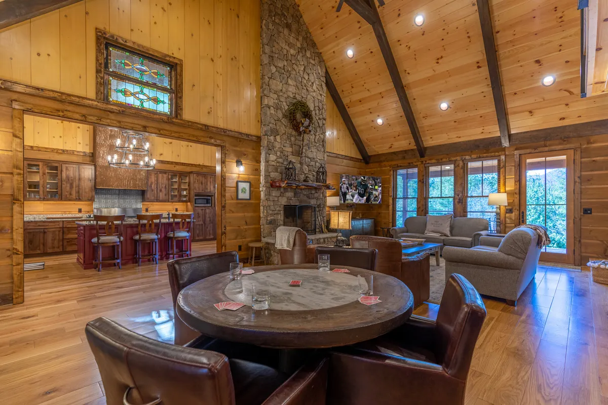 A spacious, inviting interior with a large stone fireplace as the focal point, flanked by a stained glass window and rustic wooden beam ceiling. A circular dining table sits nearby, creating a cozy space for family gatherings, meals, and game nights.
