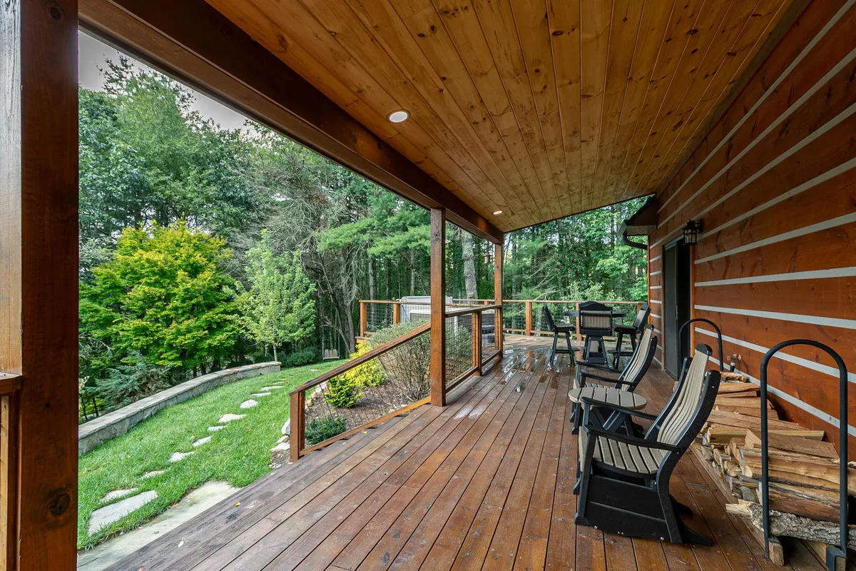 A wooden deck with comfortable chairs overlooks lush greenery and a pathway that leads to a garden. The setting is designed for relaxation, allowing both people and pets to enjoy the outdoors in a serene, mountain environment.