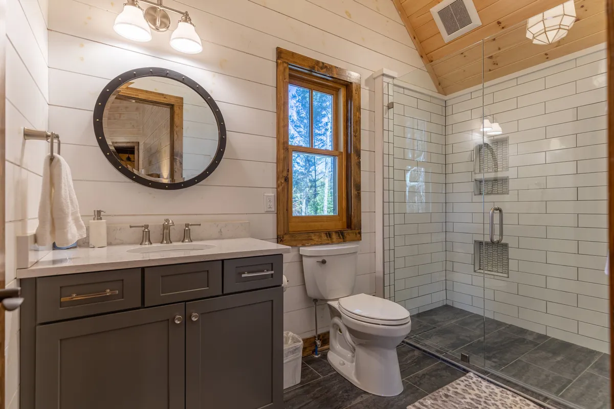 A calm and serene bathroom with a circular mirror, vanity with sink, enclosed shower, and a window letting in gentle natural light—an ideal spot for peaceful morning routines or evening unwinding.