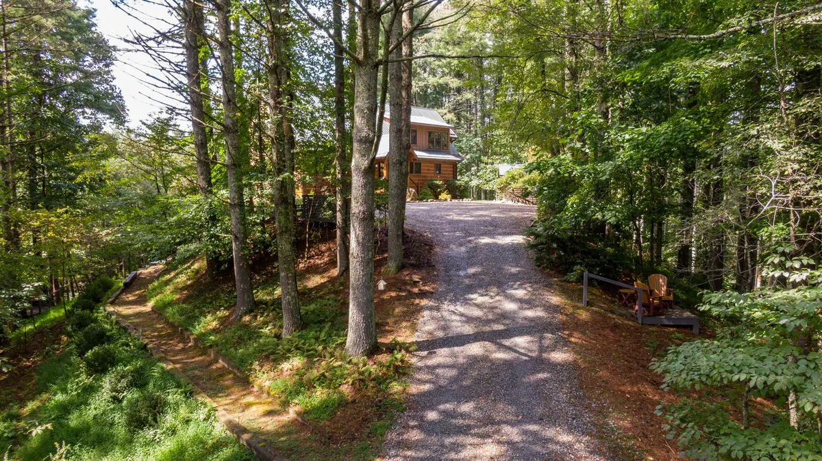 A welcoming exterior view of the house, nestled among mature trees, with a gravel driveway and a cozy seating area. The setting exudes tranquility and invites guests to step outdoors and enjoy the mountain air, while the dappled sunlight filtering through the trees creates a sense of retreat.