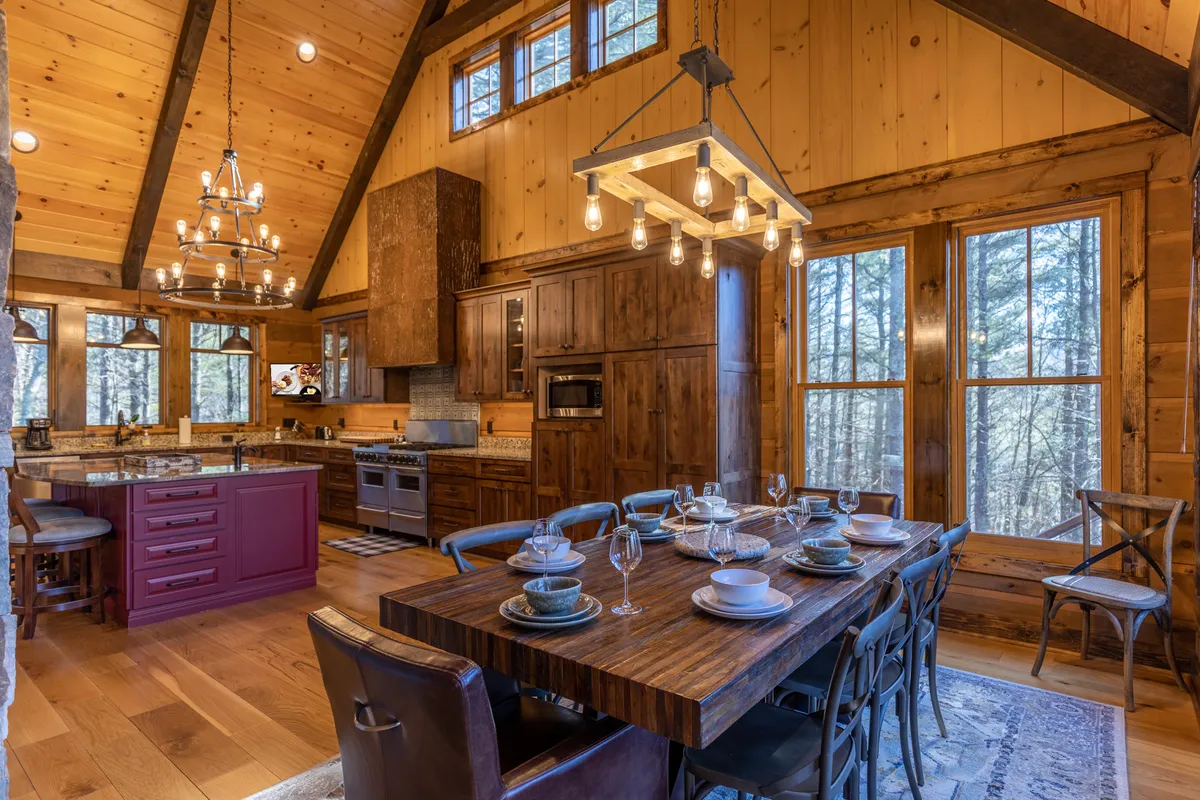A bright, airy dining area featuring a large wooden dining table, rustic kitchen cabinets, chandelier lighting, and expansive windows framing a forest view. The setup invites both shared meals and relaxed moments with loved ones.